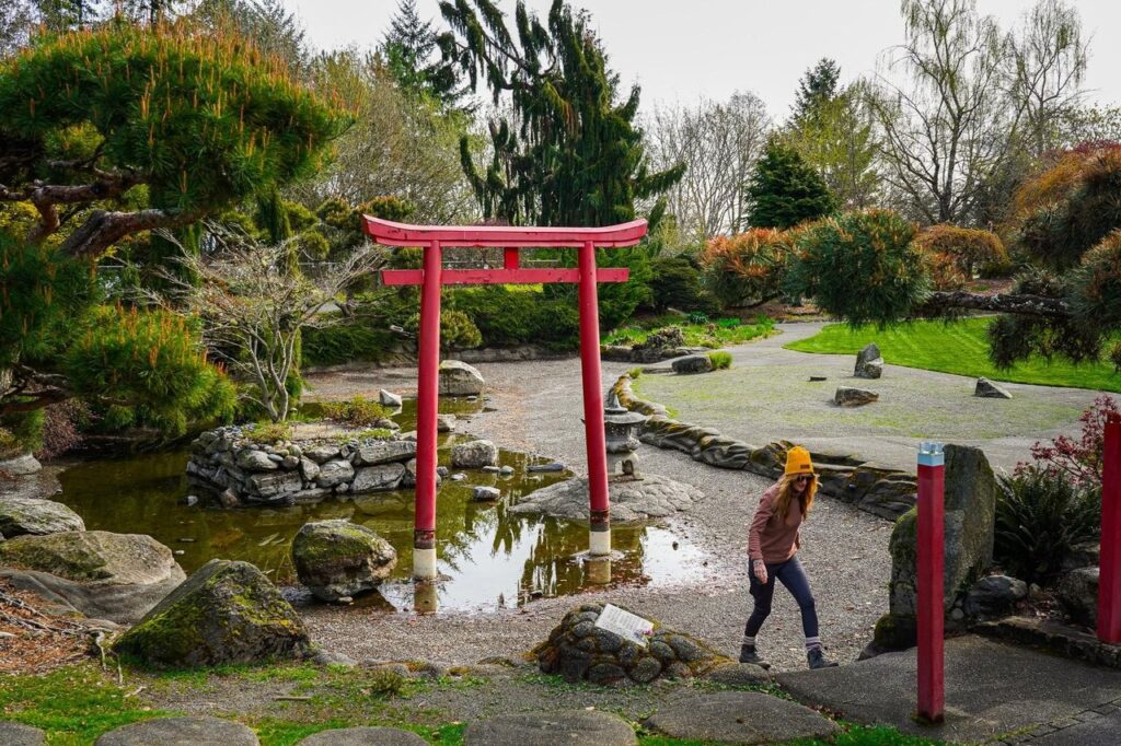tacoma-point-defiance-park-2 - junglecity.com Red torii gate over a small pond in a Japanese-style garden, with a woman in a yellow beanie walking nearby.
