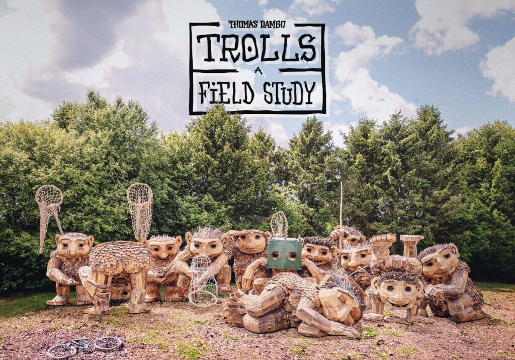 Group of carved wooden troll statues in a yard with a sign reading 'Trolls A Field Study' above.