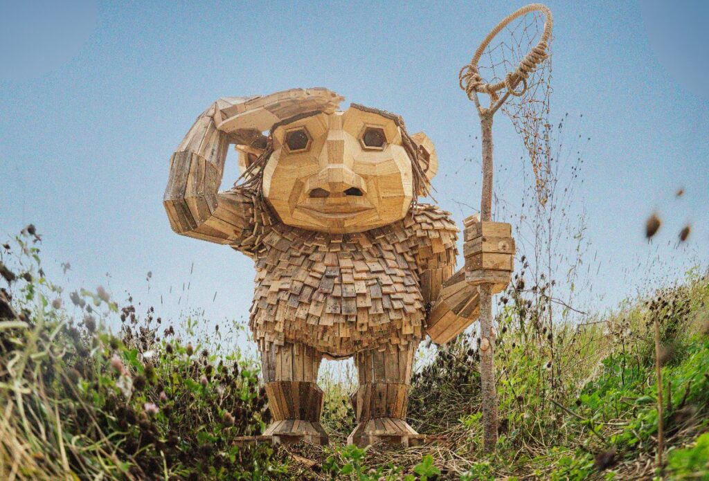 Life-size wooden sculpture of a friendly creature standing in a grassy field, holding a tall stick with a circular net.