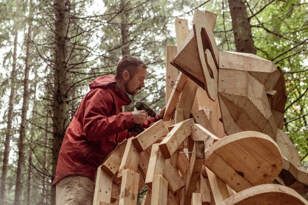 Man in a red jacket drills or sands a large wooden sculpture made of blocks in a forest setting