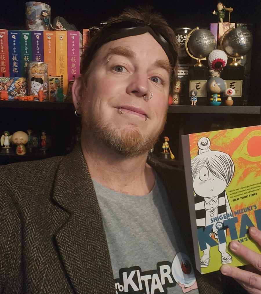 yokai-zack-1 - junglecity.com - junglecity.com Person smiling at the camera while holding a colorful illustrated book cover in the foreground, shelves of figurines in the background.