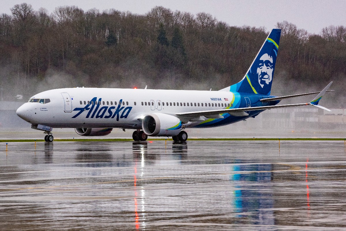 Alaska Airlines jet on a wet runway during rain, with the Alaska logo on the fuselage and a blue-green tail art reflected on the tarmac under overcast skies.