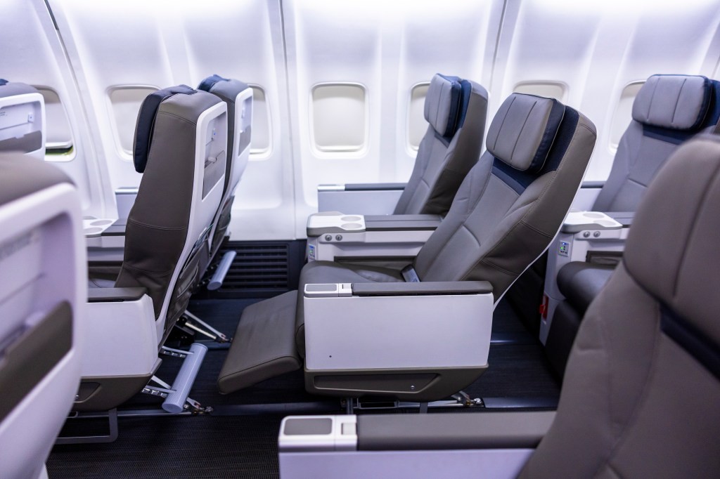 Row of gray airplane seats with wide armrests and built-in tray tables in a well-lit cabin.