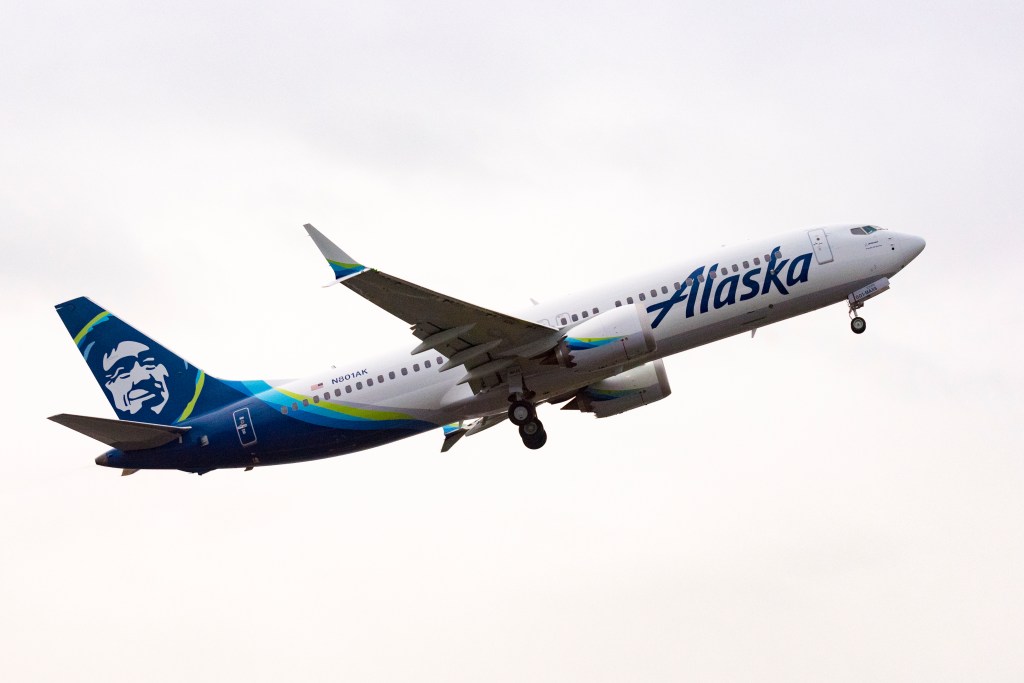 Alaska Airlines jet taking off, white fuselage with Alaska branding and blue tail featuring the logo against a pale sky.