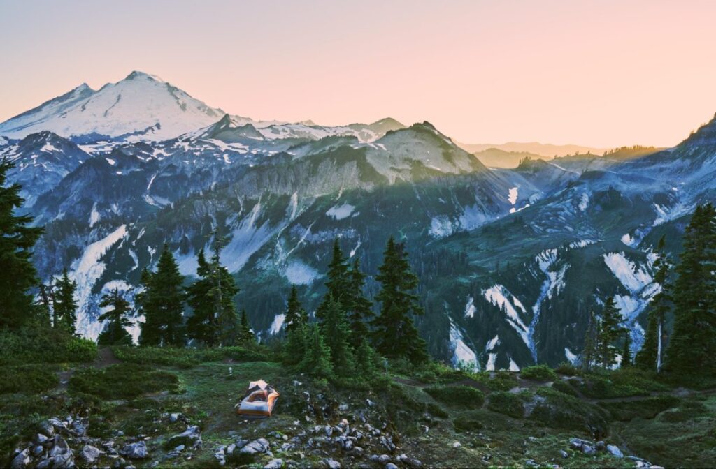 artist-point-2 - junglecity.com Orange tent pitched on a rocky alpine meadow with evergreen trees, a backdrop of snow-capped mountains at sunset/ sunrise.