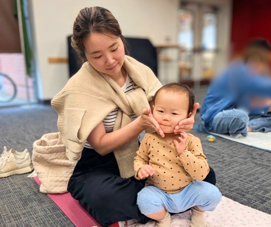 Woman sits on a pink mat indoors, gently pinching a smiling toddler's cheeks.