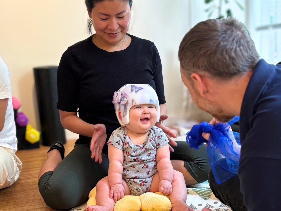 Happy baby wearing a soft protective helmet sits on a mat, supported by a caregiver while a clinician shows a blue fabric indoors.