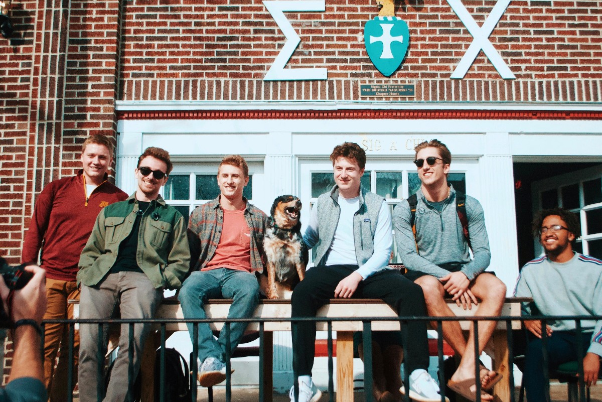 Six young men and a dog sit on a railing outside a brick fraternity house with Greek letters on the wall behind them.