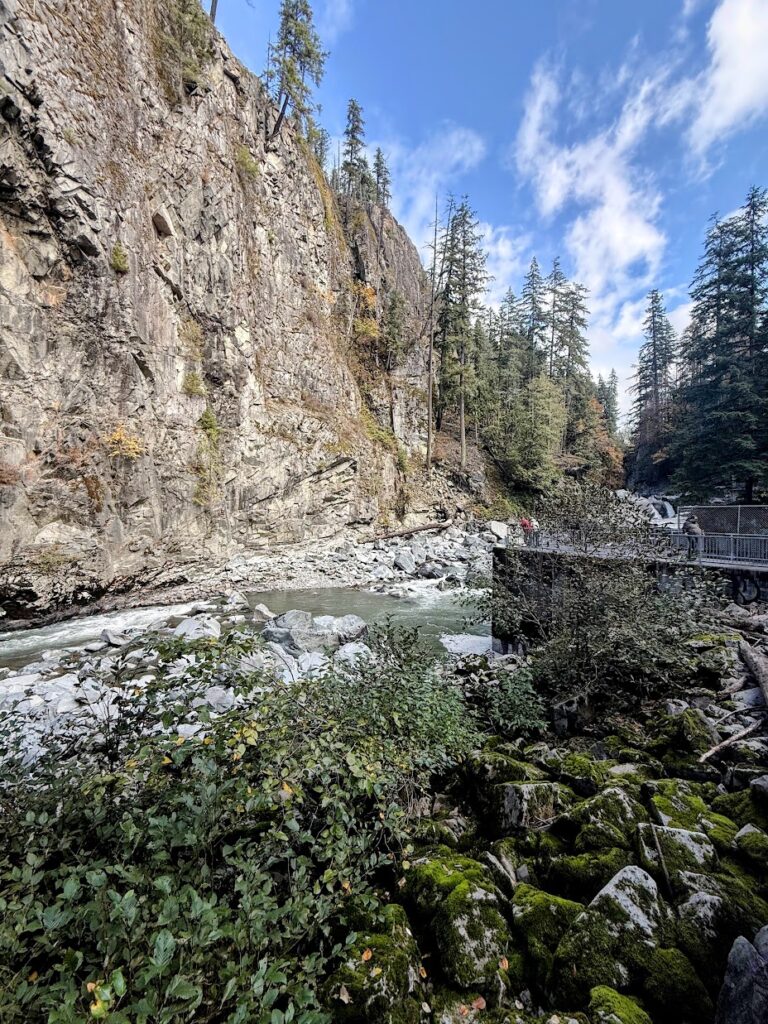 granite-falls-fish-ladder-2 - junglecity.com Steep rocky cliff beside a rushing river, with evergreen trees and a pedestrian overlook where people stand on a railing.