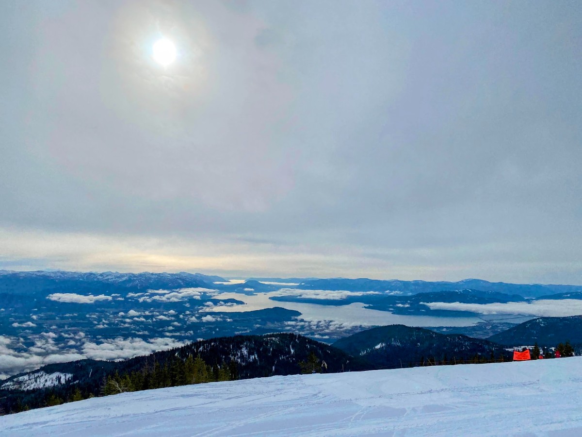 Snow-covered mountain slope overlooking a bay with islands, under a hazy sun and cloudy sky.