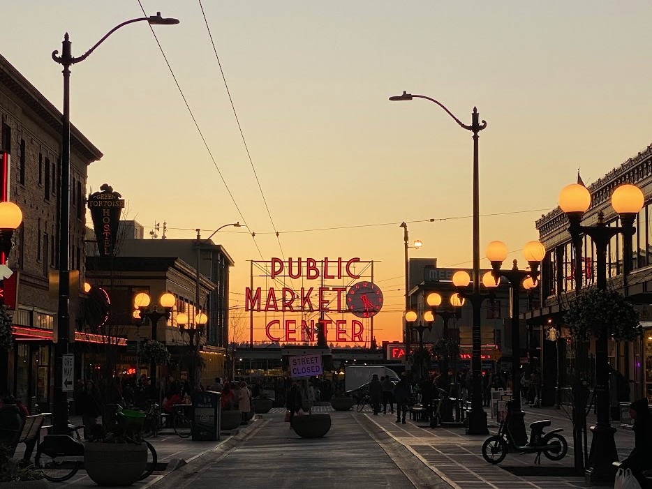 Sunset street scene toward a large red neon sign that reads 'Public Market Center', with lamp posts and people walking along a pedestrian plaza.