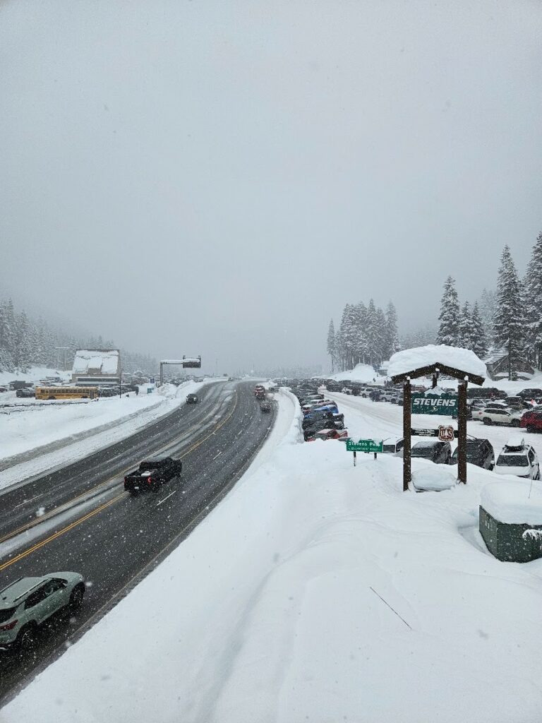 stevens-pass-5 - junglecity.com Snowy mountain highway with cars traveling and parked along the snow-packed shoulder, pine trees in the distance and a Stevens sign near the road.