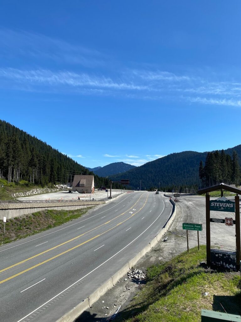 stevens-pass-6 - junglecity.com Winding mountain highway through a forested valley with a small building on the left and a Stevens Pass sign on the right under a bright blue sky