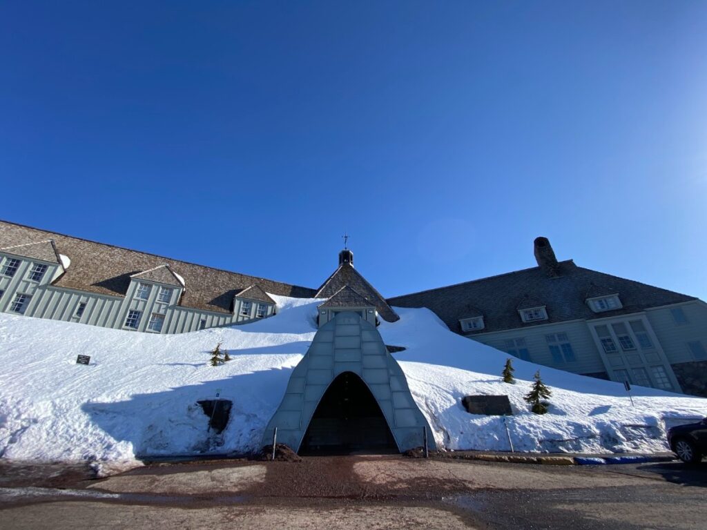 Snow-covered hillside leads to a central arched glass tunnel entrance of a mountain-style lodge against a clear blue sky.